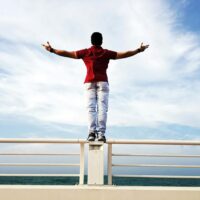 A person standing on a railing by the sea with arms outstretched under a clear sky, symbolizing freedom.