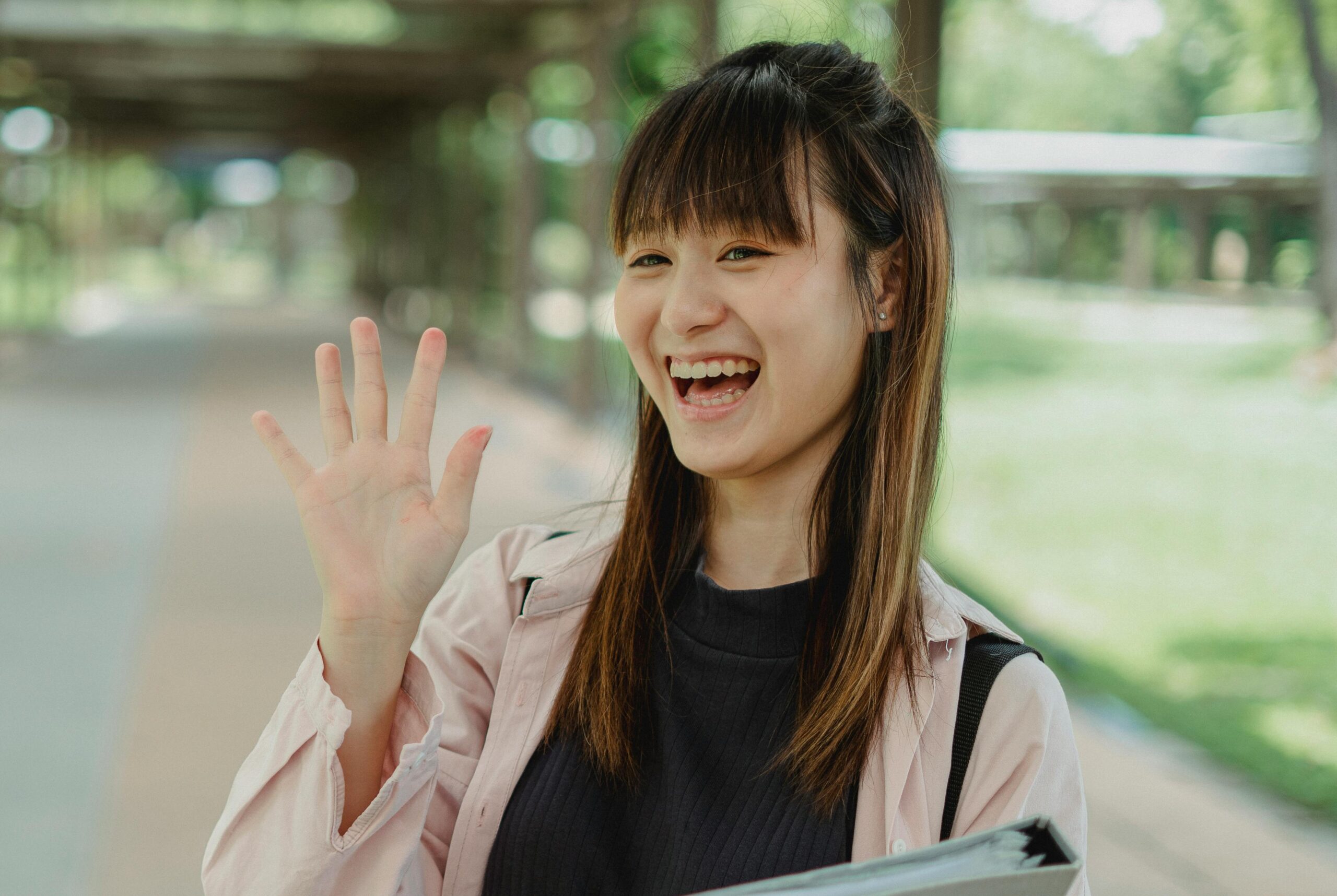 Smiling woman waving hello while standing outdoors on a sunny day.